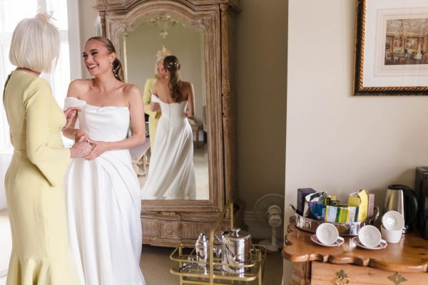 A bride in a white dress holds hands with a woman in yellow, both smiling in a room at Fawsley Hall, with a mirror, tea set, and various refreshments visible—a perfect moment for Fawsley Hall photography.