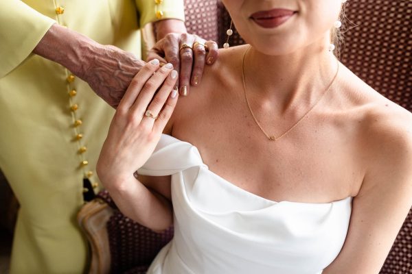A bride in a white dress sits as an older woman in yellow rests her hand on the bride’s shoulder. Both women wear rings and have manicured nails—an elegant moment captured by Fawsley Hall photography.